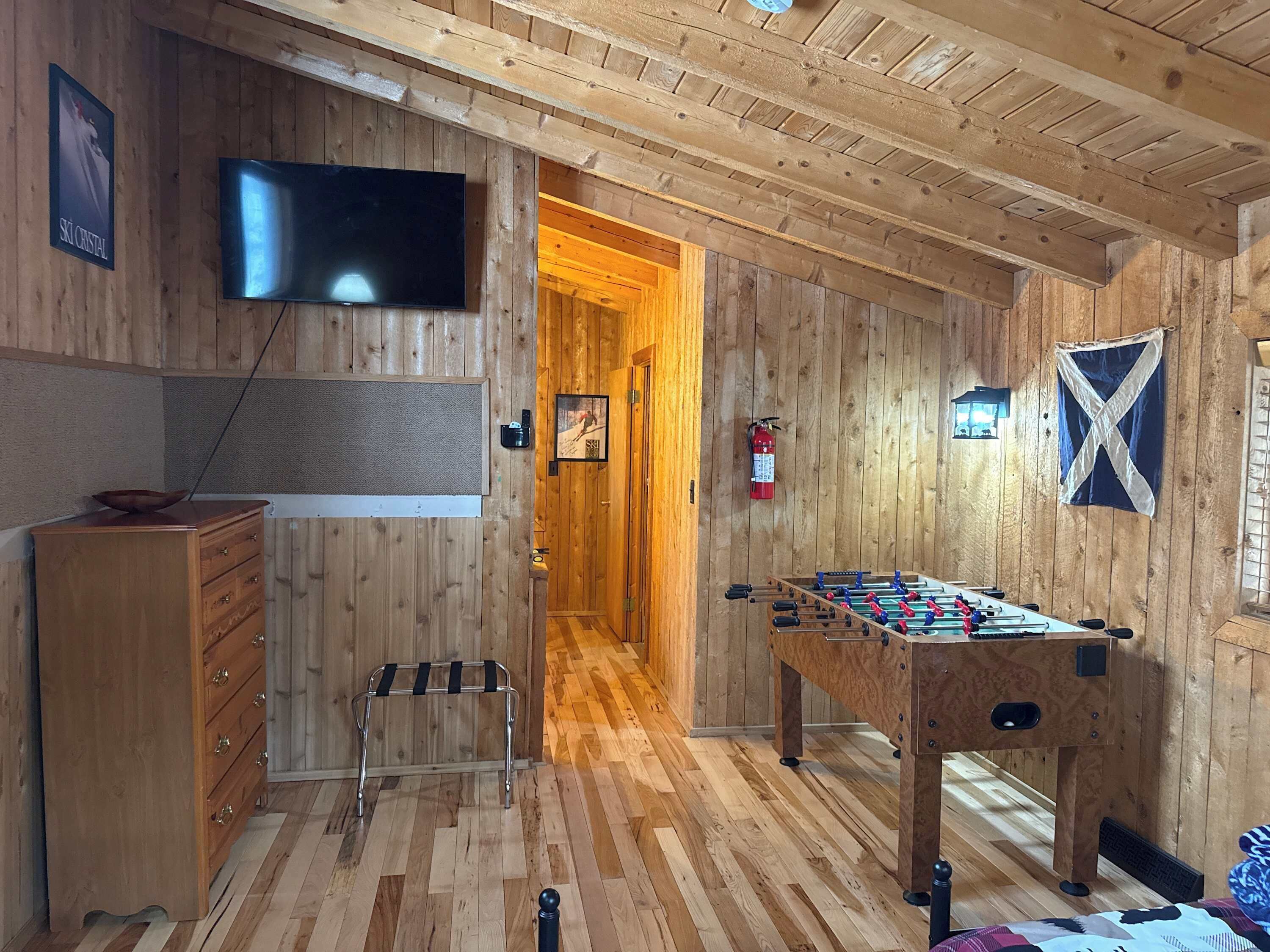 Une pièce au plancher de bois, équipée d’un baby-foot, d’une télévision, d’une commode en bois et d’une porte donnant sur un autre espace.