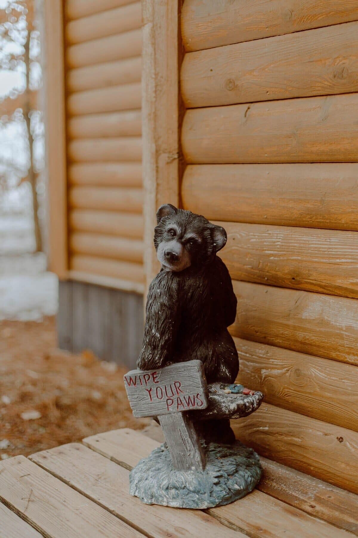 A wooden bear figurine sitting on a bench with a sign that reads "Wipe Your Paws".
