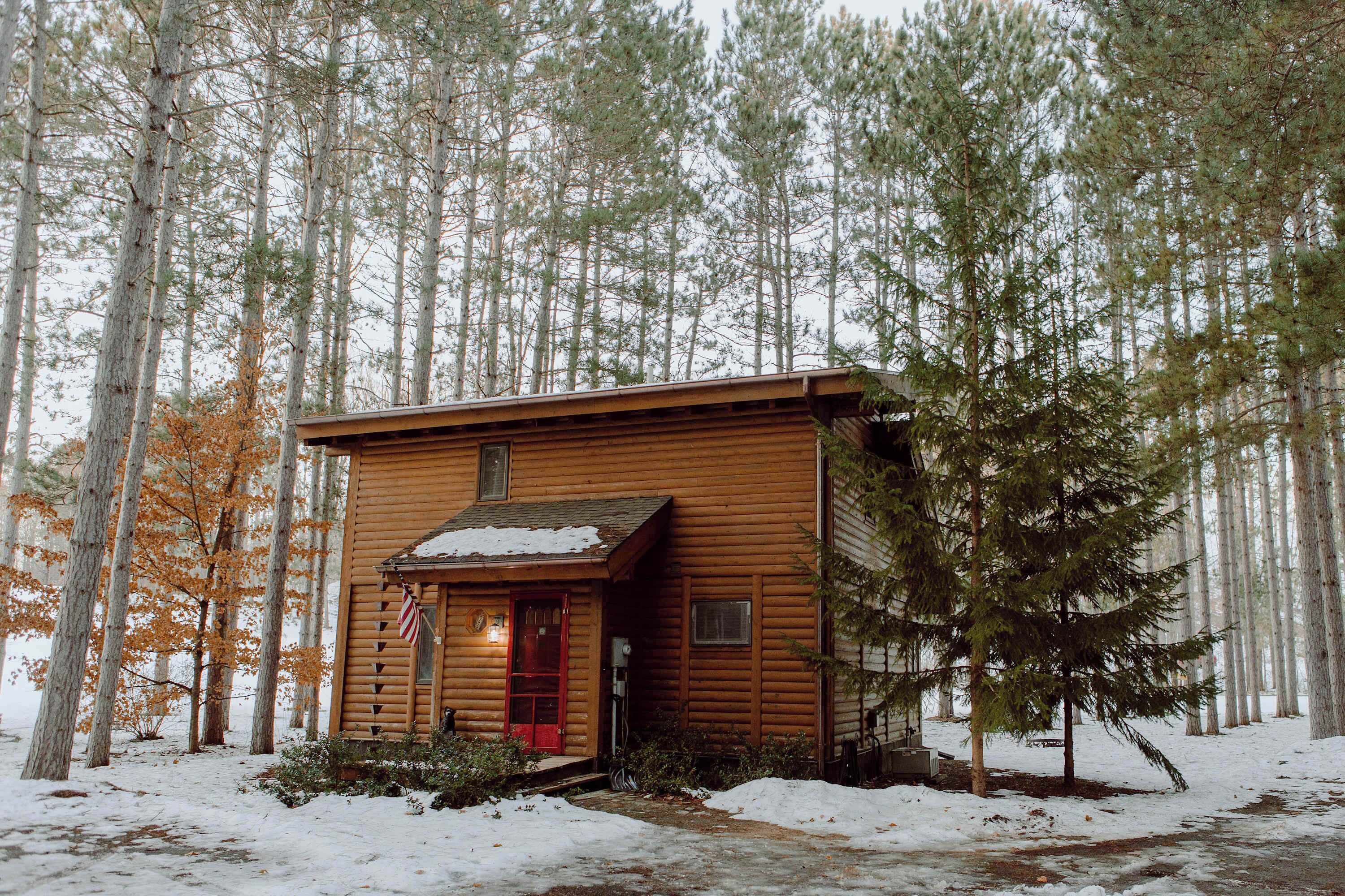 A log cabin with a red door, surrounded by tall pine trees and covered in snow.