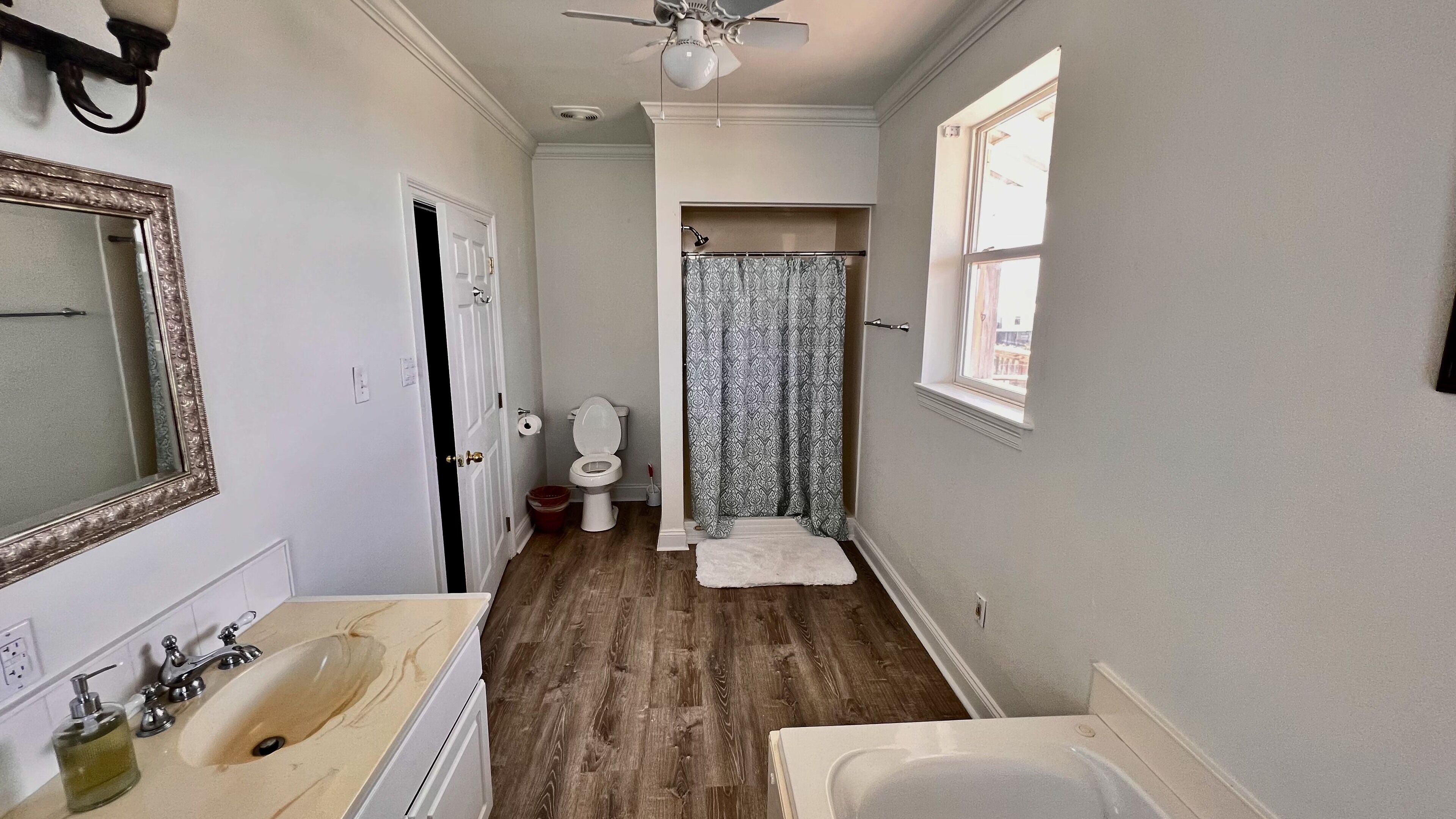 A narrow bathroom with a wooden floor, a white sink, a mirror, and a shower with a patterned curtain.