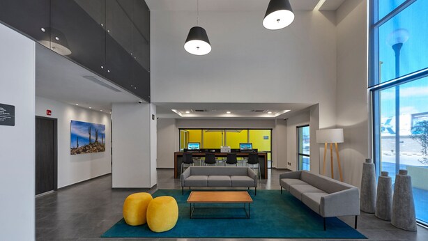 Um lobby moderno com sofá, banquinho e mesa de centro, uma mesa de escritório com computadores e grandes janelas.