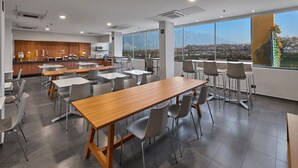 Uma sala de jantar moderna com uma longa mesa de madeira e cadeiras cinzentas, amplas janelas com vista para montanhas, e uma área de cozinha com armários de madeira e micro-ondas.