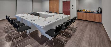 Uma sala de conferências com uma mesa comprida, cadeiras pretas, uma tela de projetor, uma porta de madeira e uma área de kitchenette.