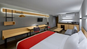 Quarto de hotel moderno com uma cama grande, uma escrivaninha, uma cadeira e uma cozinha compacta.