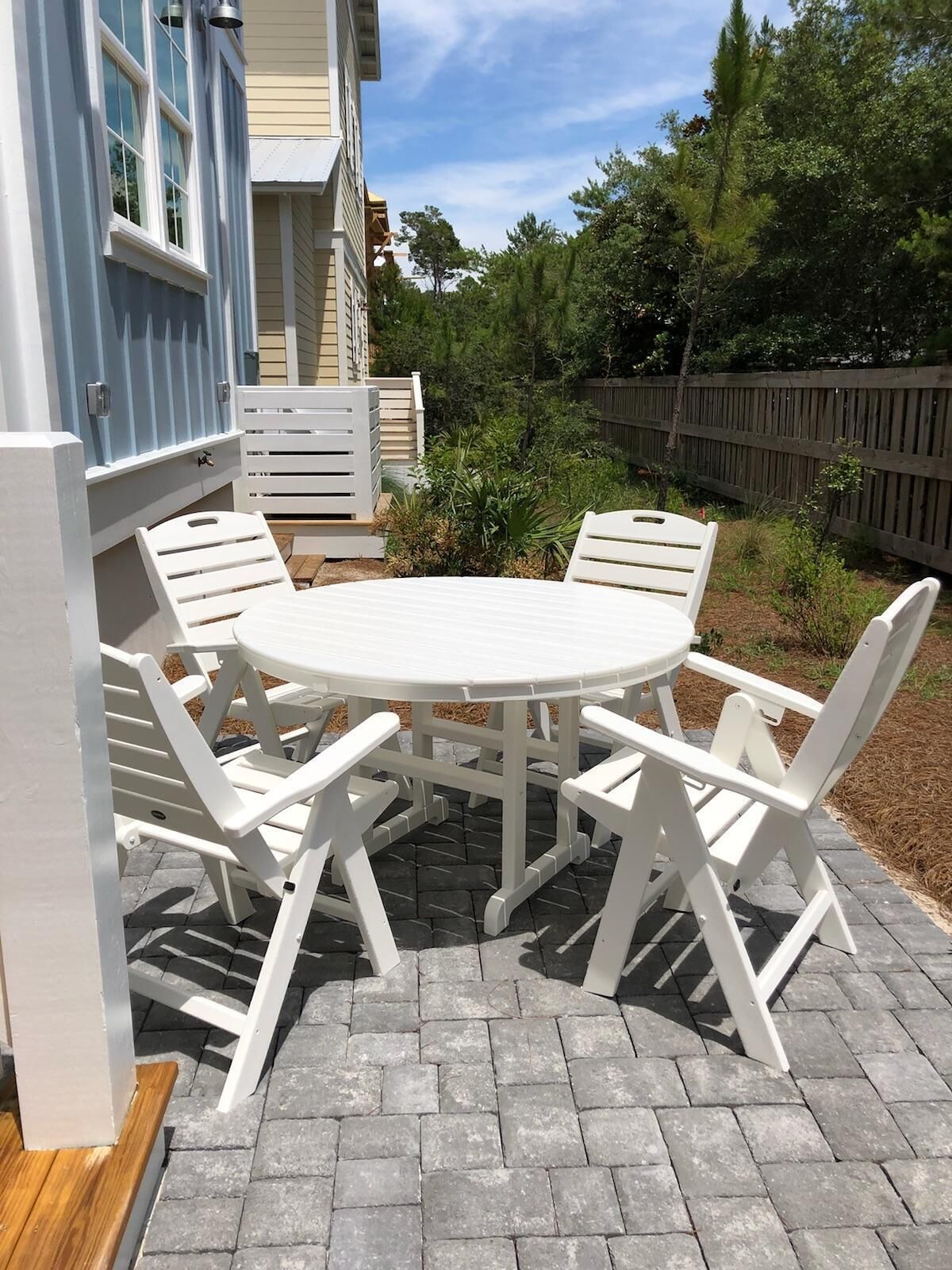 A patio with a round table and white chairs, surrounded by a wooden fence and greenery.