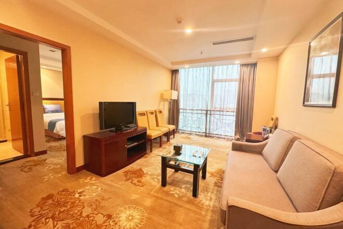 Une chambre d’hôtel équipée d’un canapé, d’une télévision, d’une table basse, d’un fauteuil et d’une grande fenêtre avec des rideaux.