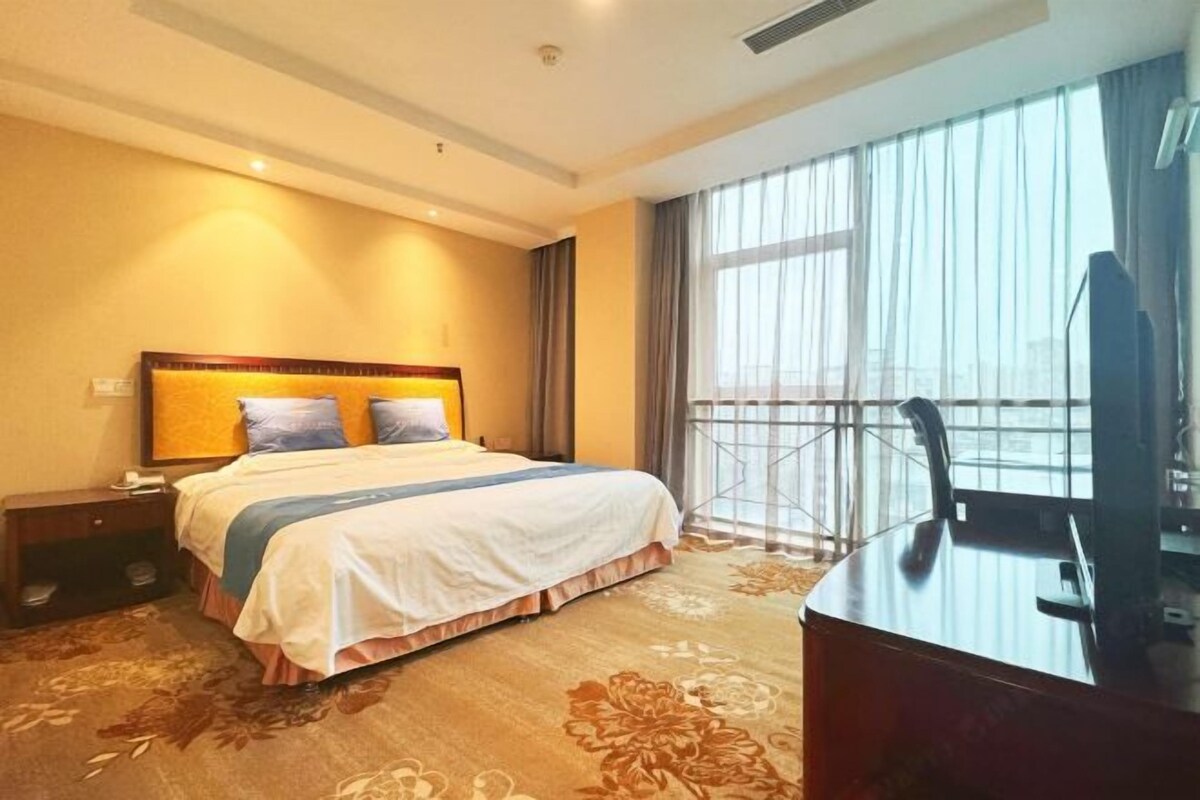 A hotel room with a large bed, a desk, and a view of the city through large windows.