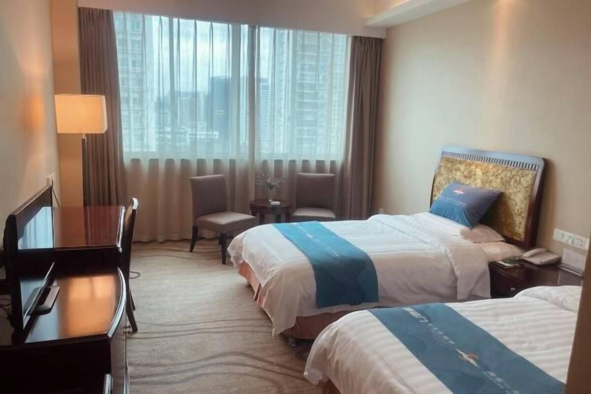 Une chambre d’hôtel avec deux lits, un bureau, une chaise, une petite table et une vue sur les bâtiments à travers de grandes fenêtres.