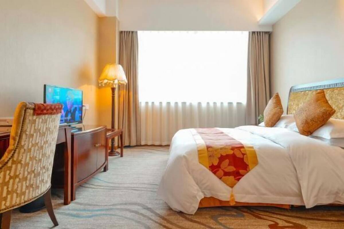 Une chambre d’hôtel avec un lit, un bureau, une chaise, une lampe et une fenêtre avec des rideaux.