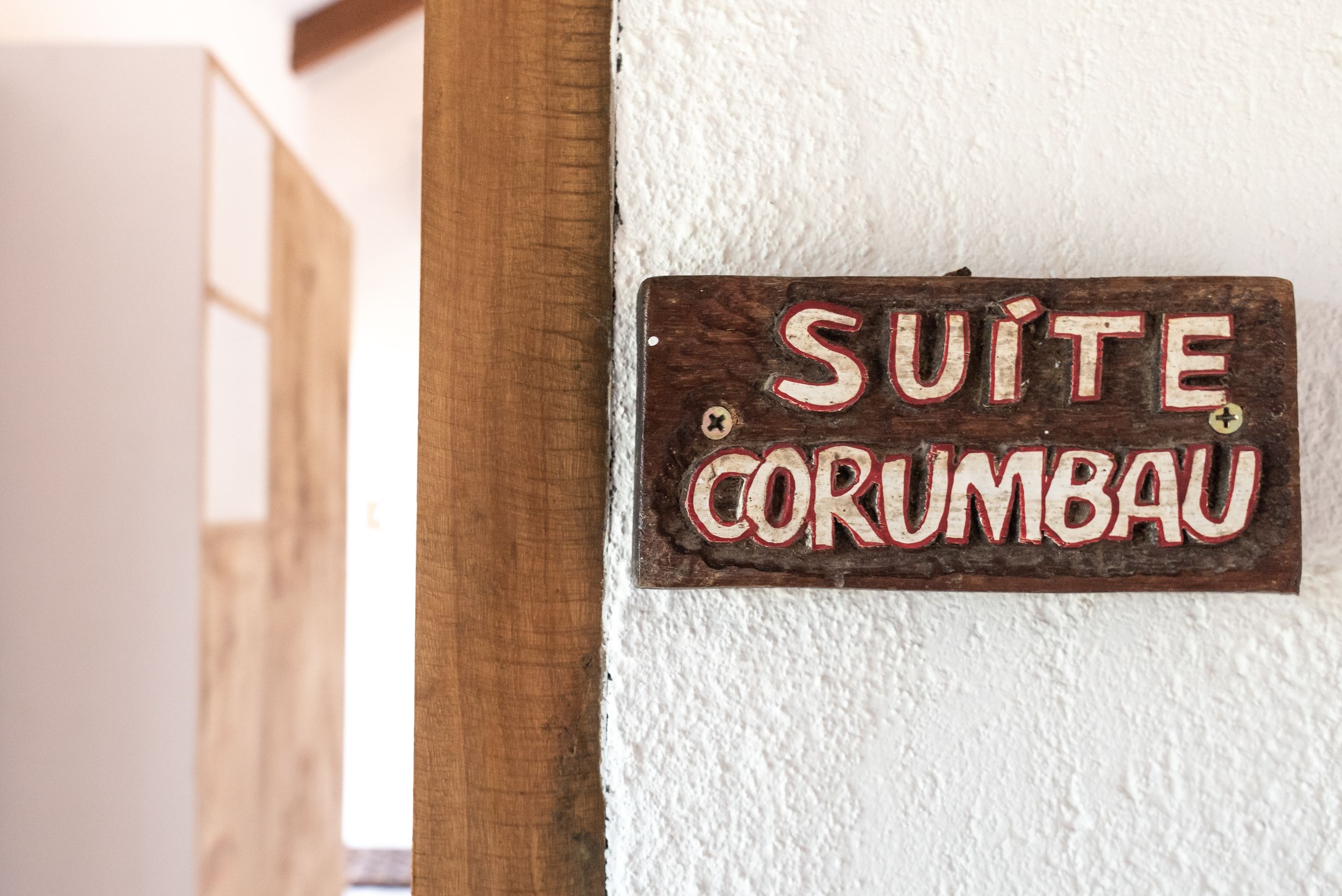 Un letrero de madera con el texto "SUITE CORUMBAU" fijado a una pared blanca.