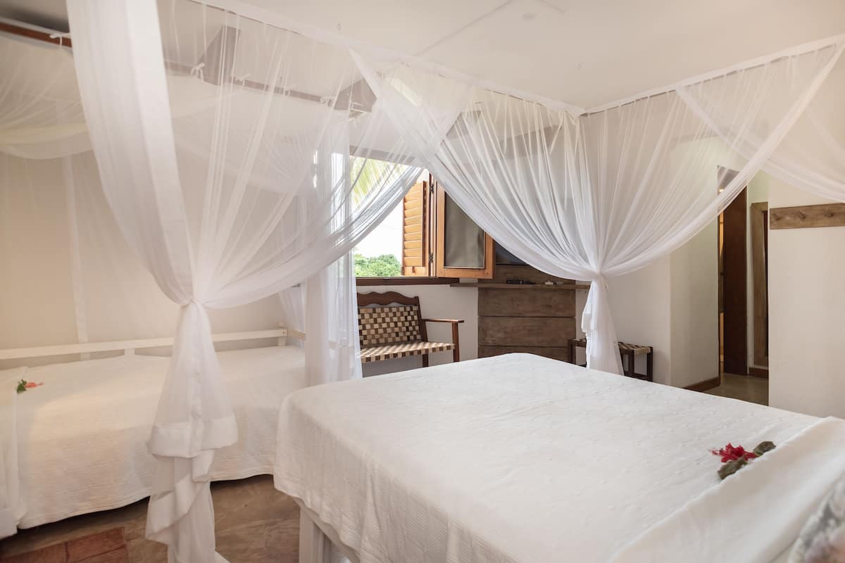 Een slaapkamer met twee bedden, elk voorzien van witte beddengoed en een hemelbed. Er staat een houten bankje en een tafeltje in de kamer.