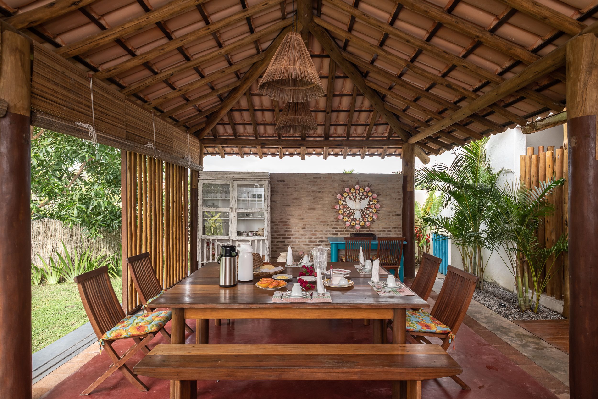 A covered dining area with a wooden table set for a meal, surrounded by chairs with colorful cushions.