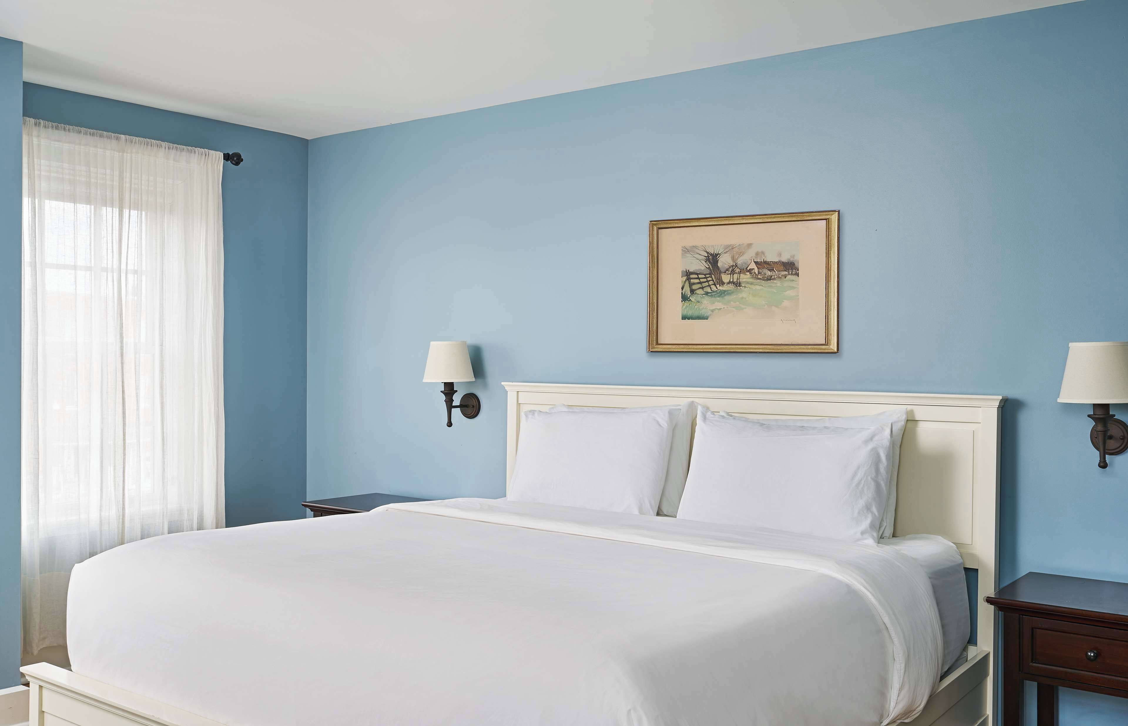 A neatly made bed in a room with light blue walls, a framed picture on the wall, and two bedside lamps.