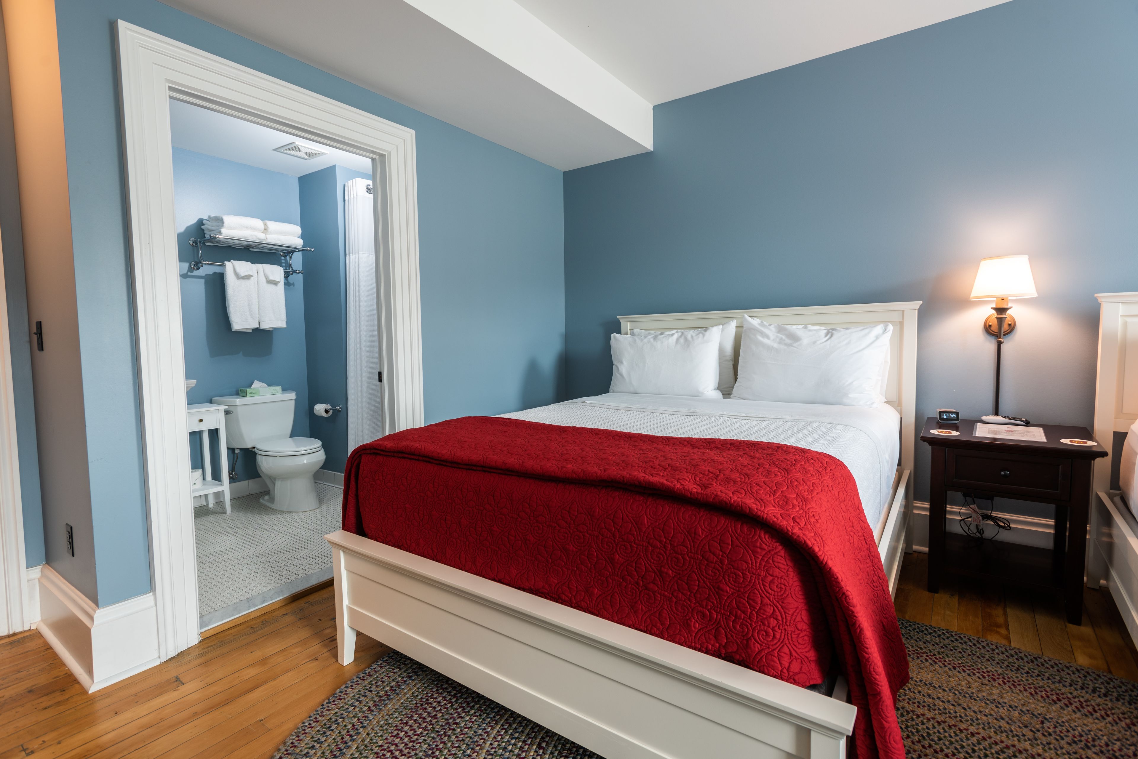 A bedroom with a bed, a nightstand, a lamp, and a bathroom visible through an open door.