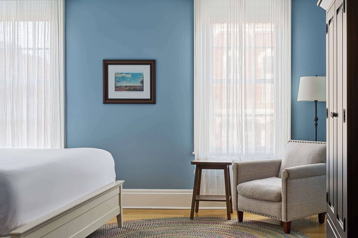 A bedroom with a bed, a chair, a small table, a framed picture on the wall, and a lamp.