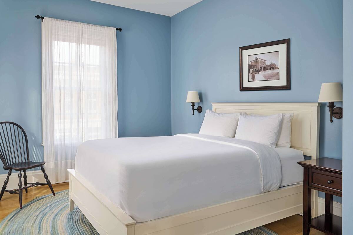 A bedroom with a bed, a chair, a nightstand, a lamp, and a framed picture on the wall.
