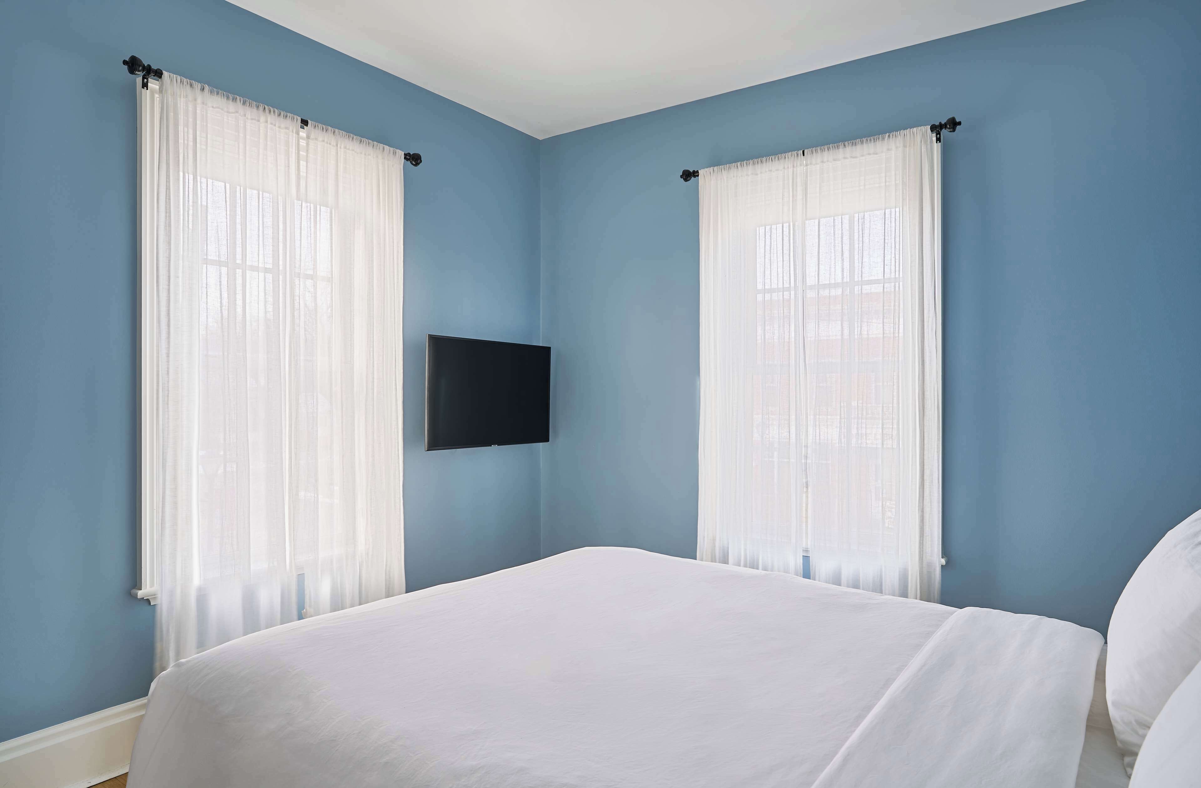 A bedroom with a bed, two windows with sheer curtains, and a flat-screen TV mounted on the wall.
