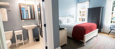 A bedroom with a bed, a nightstand, a dresser, a window with curtains, and a view of buildings outside.