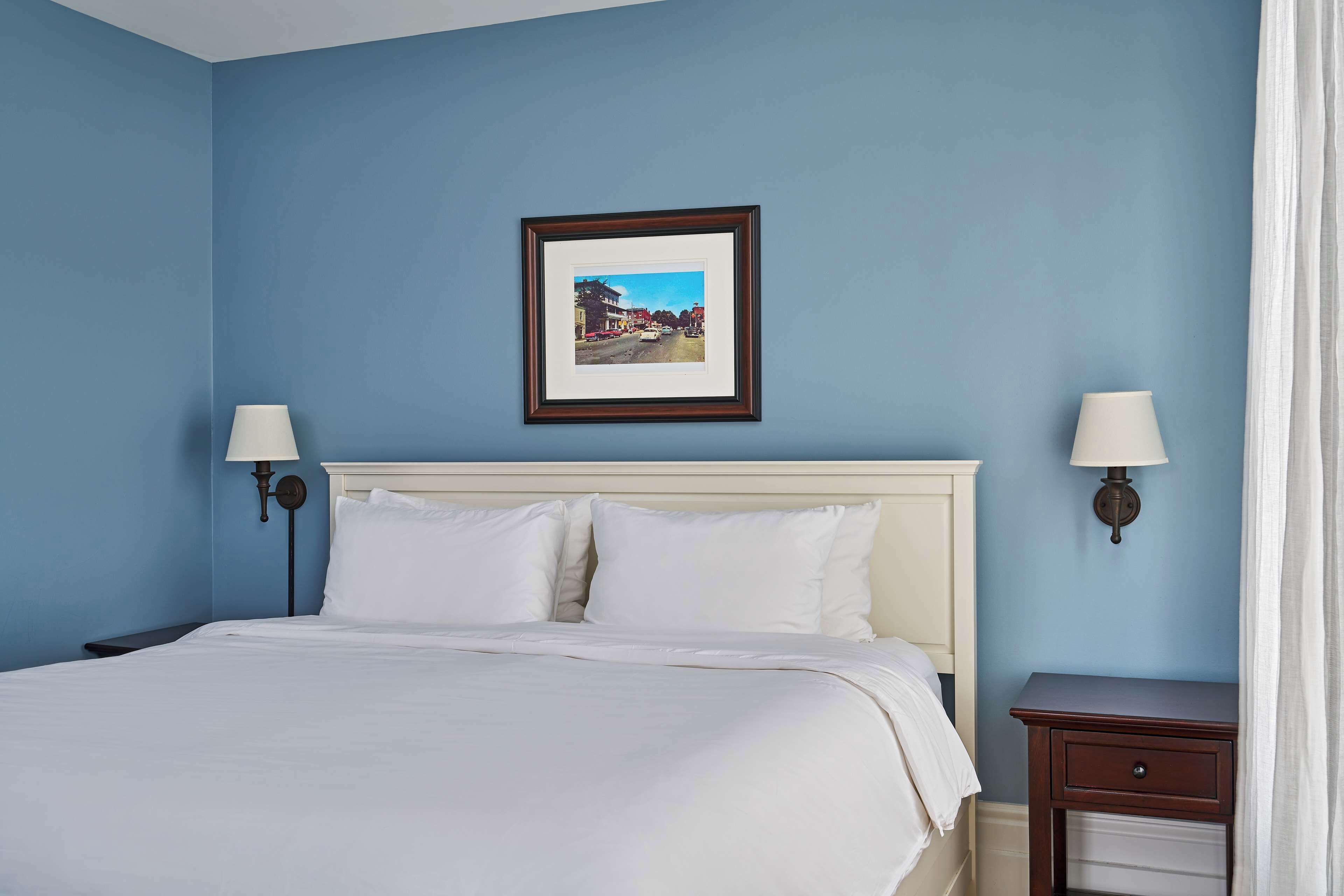 A hotel room with a large bed, two bedside tables with lamps, a framed picture on the wall, and a nightstand with a drawer.