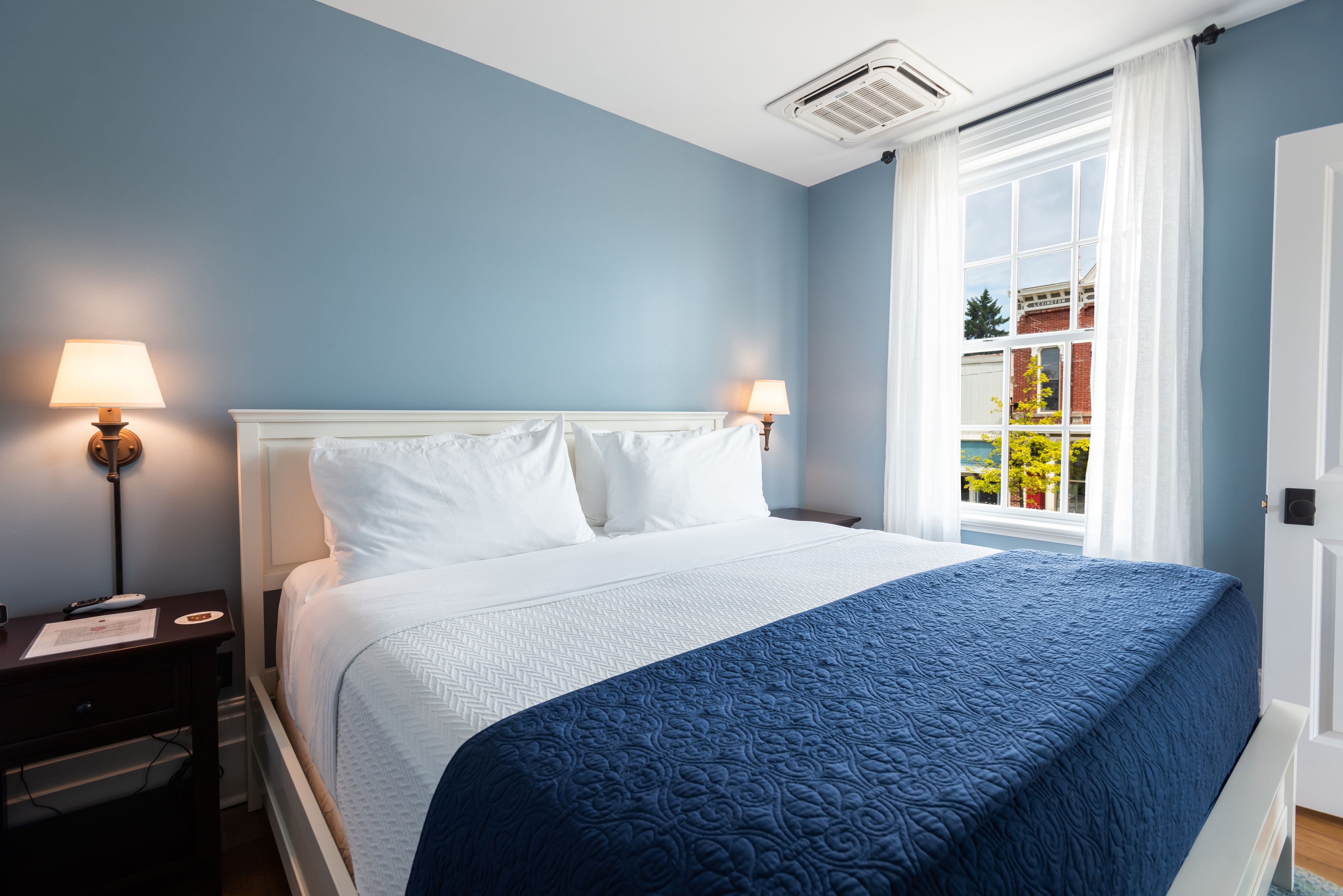 Un lit bien fait, avec du linge de lit blanc et une couverture bleue, deux lampes de chevet, une table de nuit et une fenêtre ornée de rideaux blancs.