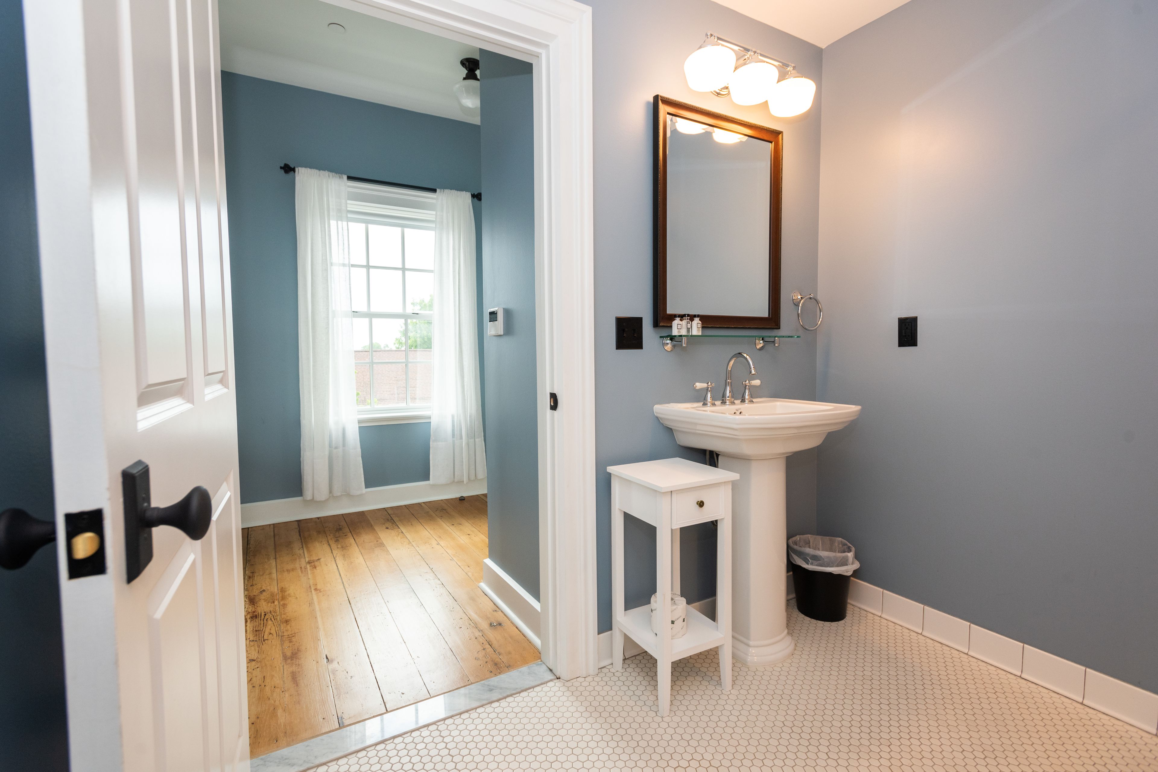 Une salle de bain avec un lavabo sur colonne blanc, un miroir avec éclairage intégré et une porte blanche donnant sur une pièce dont le sol est recouvert de parquet.