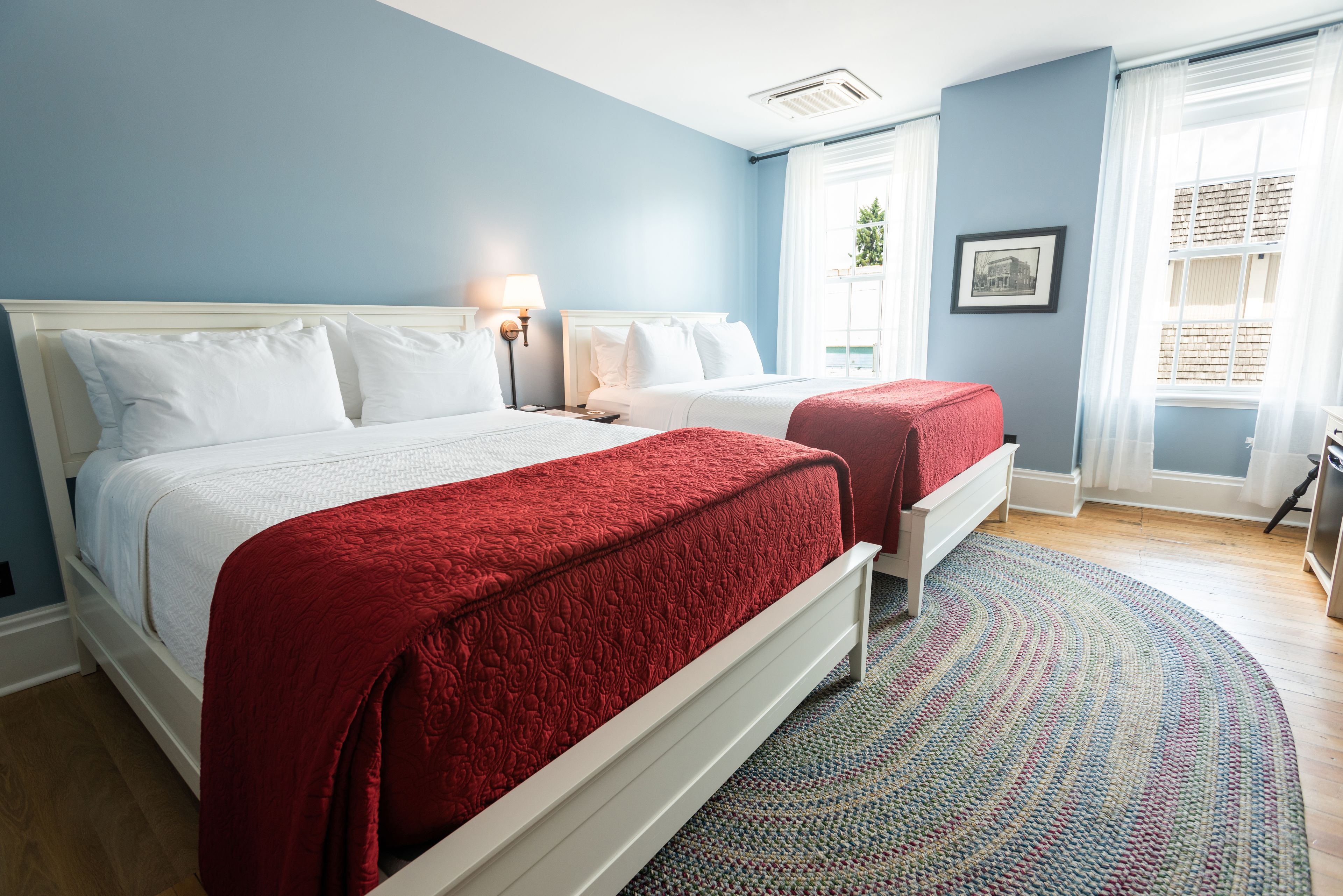 A hotel room with two beds, a large window with curtains, a framed picture on the wall, and a patterned rug.
