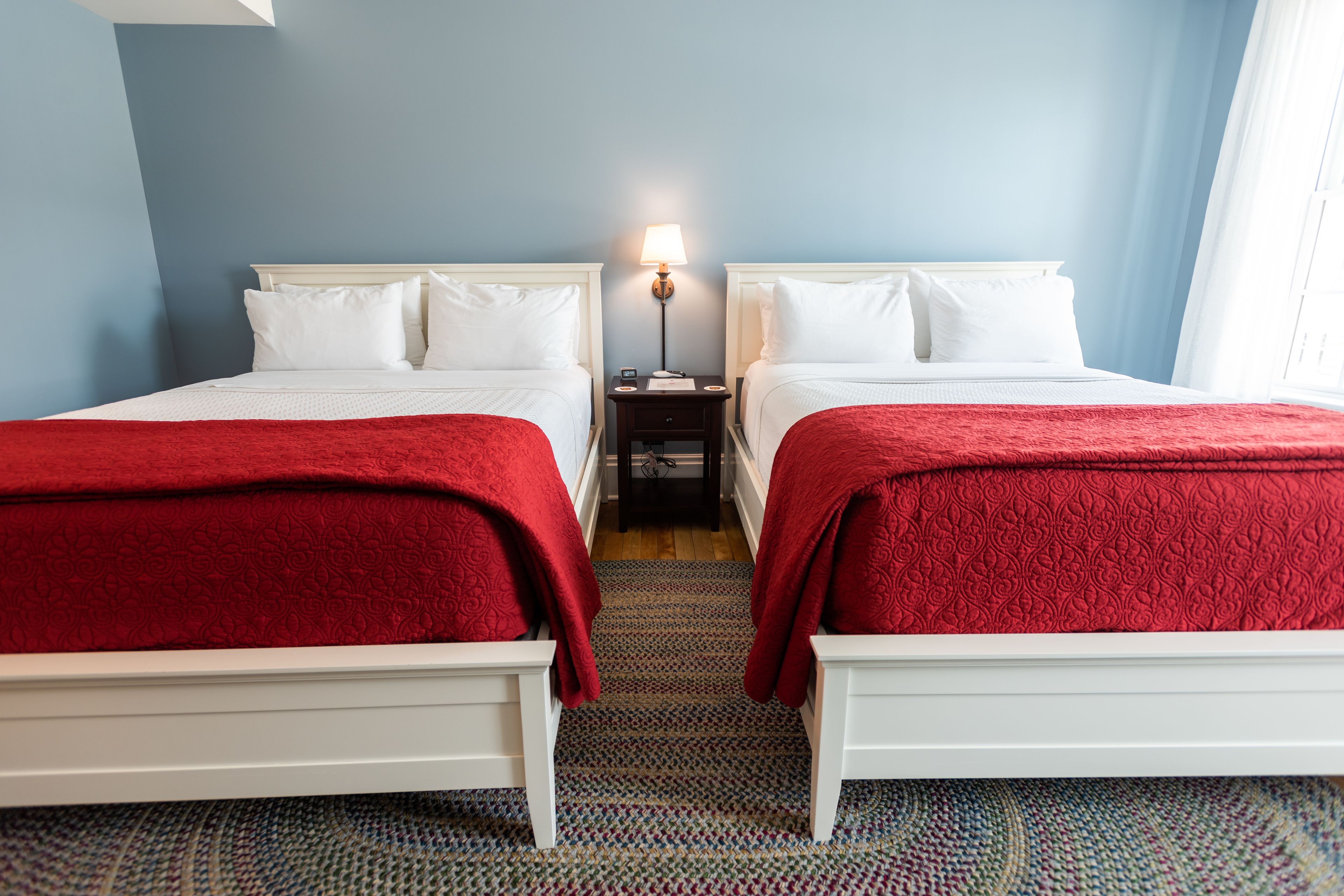 Habitación de hotel con dos camas, cada una con cabeceros blancos y colchas rojas. Hay una mesita de noche con una lámpara entre las camas. La habitación tiene una pared azul y una alfombra estampada.