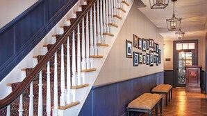 Un escalier avec des balustrades en bois et une moquette à motifs, menant à un palier où se trouvent un banc et des tableaux encadrés accrochés au mur.