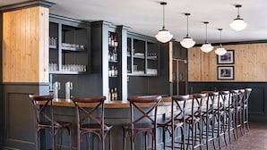 Un espace bar doté d’un long comptoir et de nombreux tabourets, d’étagères remplies de verres et de bouteilles, et d’un éclairage suspendu.