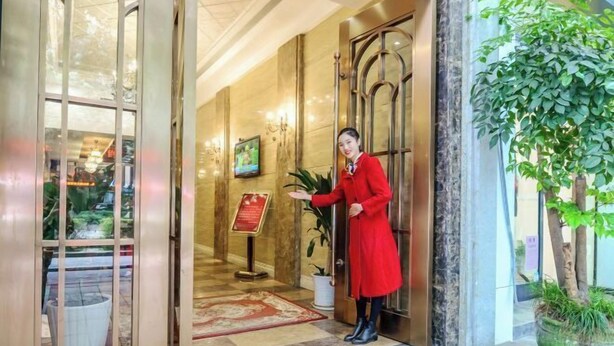 Entrada d'hotel amb un membre del personal portant roba vermella, una taula de recepció, una planta en test i un sòl de marbre.