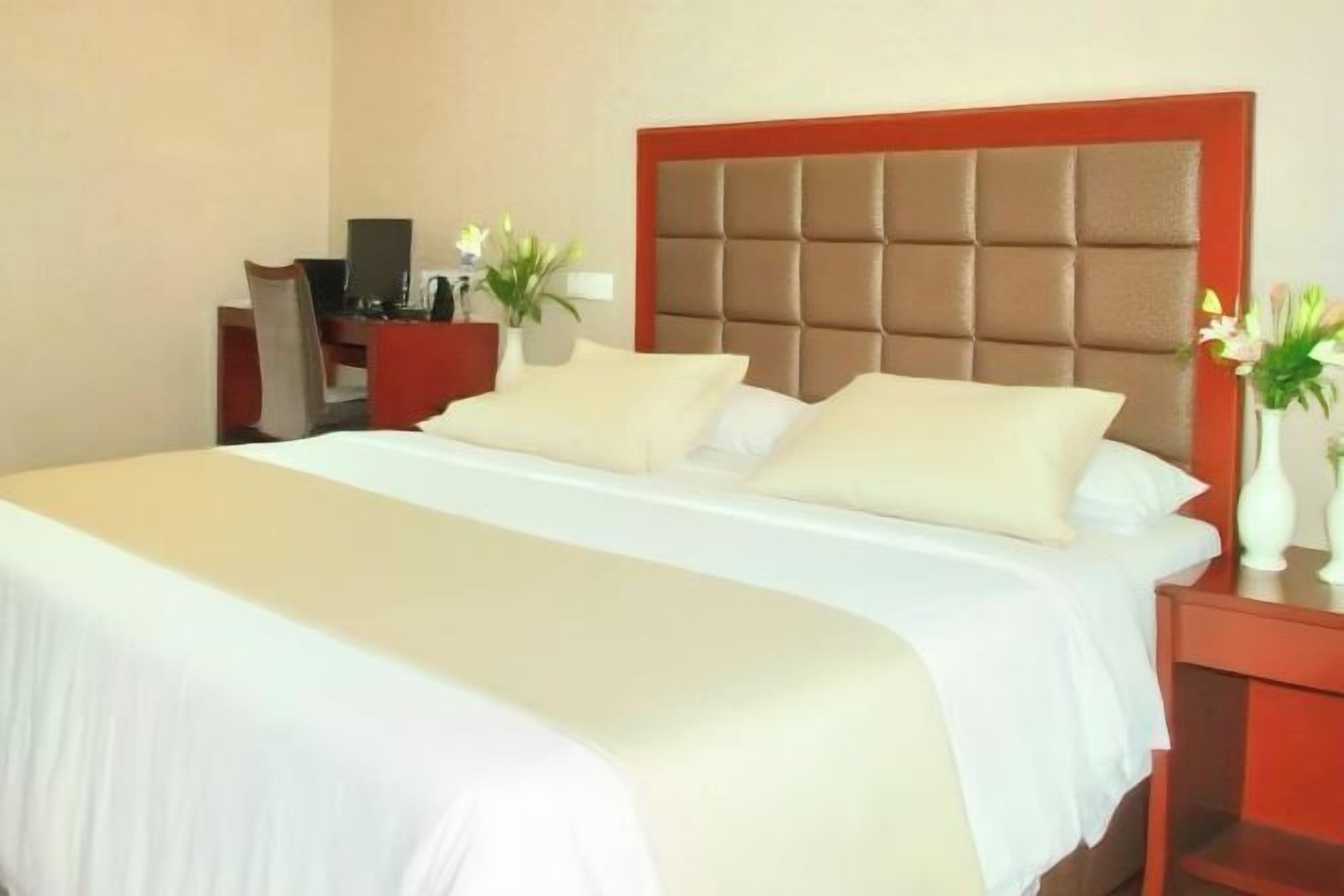 Een hotelkamer met een groot bed, een rood hoofdeinde, een bureau met stoel en een klein tafeltje met een bloemenvaas.