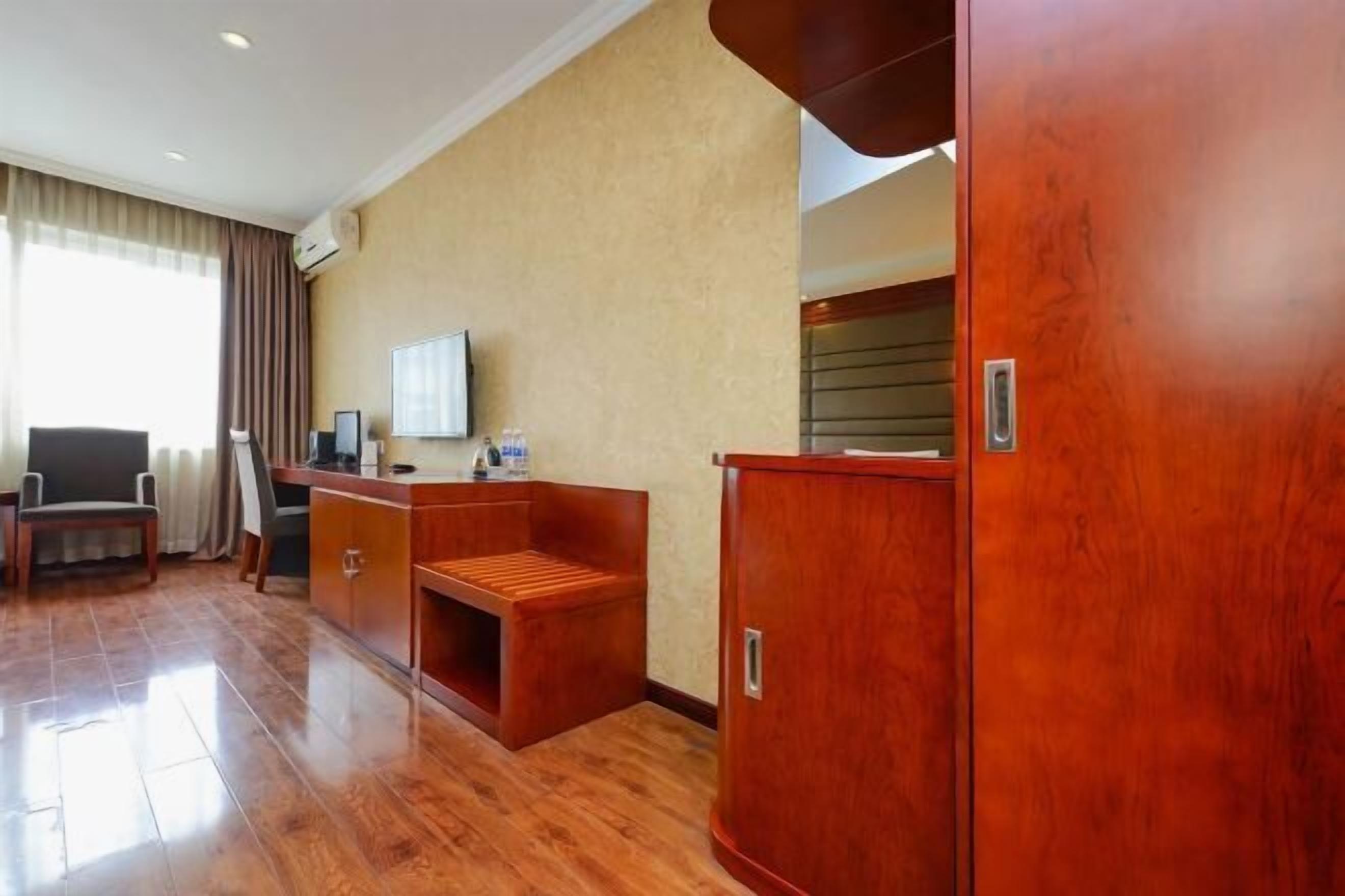 Habitación de hotel con escritorio de madera, una silla, un pequeño rincón de descanso, armario y televisor en la pared.