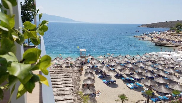 A beach with numerous umbrellas and lounge chairs, a clear view of the sea, and a mountainous backdrop.