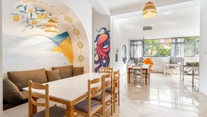 A spacious dining area with a long table and chairs, a mural on the wall, and a kitchen in the background.