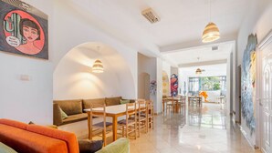 A spacious living area with a sofa, dining table, and chairs. There are paintings on the walls and a door leading to another room.