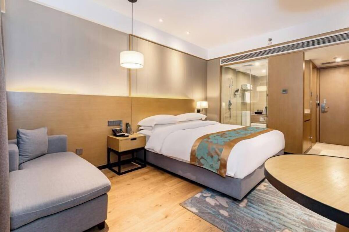 A modern hotel room with a bed, a bedside table, a lamp, a sofa, and a round table.