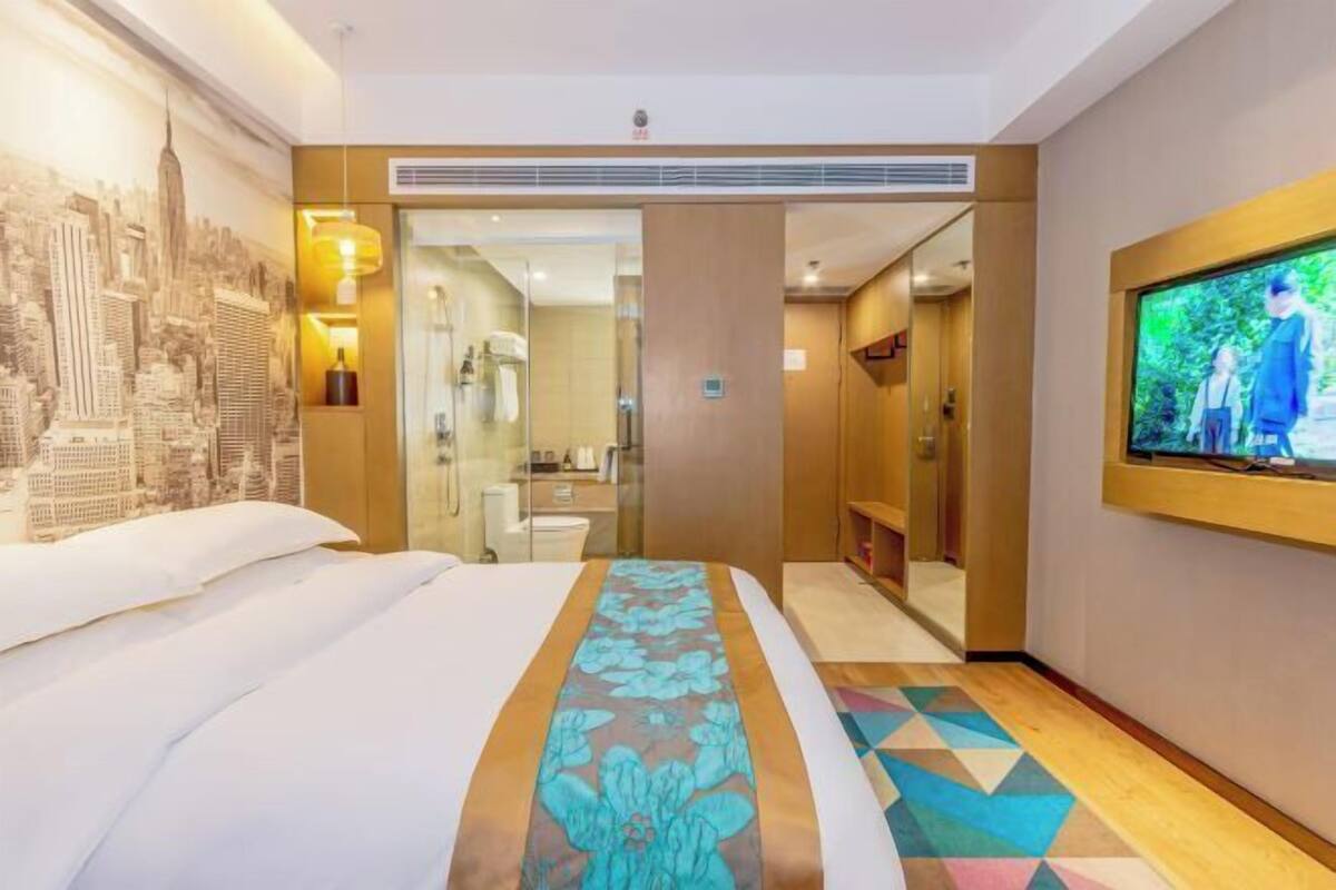 A hotel room with a bed, a cityscape mural, a television, and a bathroom visible through a glass door.