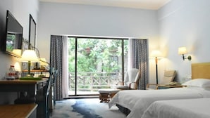 Quarto de hotel com cama, escrivaninha, cadeira, abajur e vista para o verde pela janela.