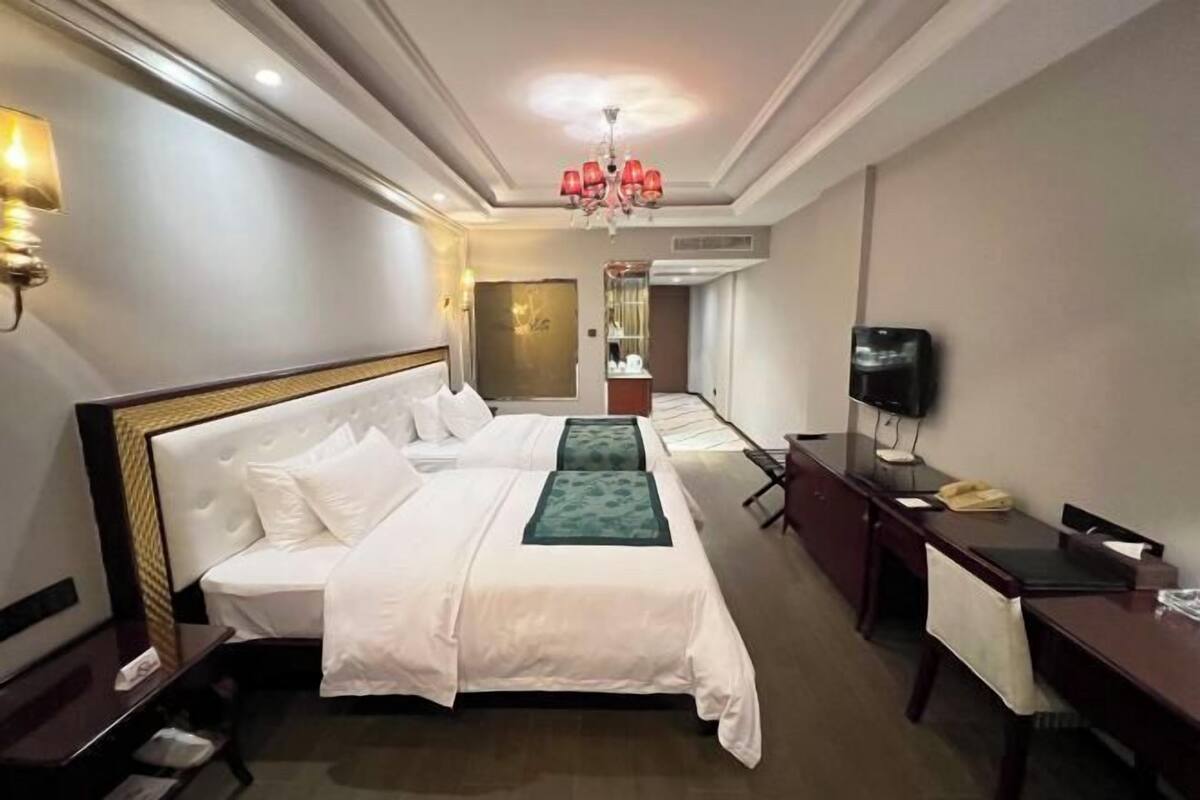 A hotel room with two beds, a desk, a chair, a TV, and a chandelier.