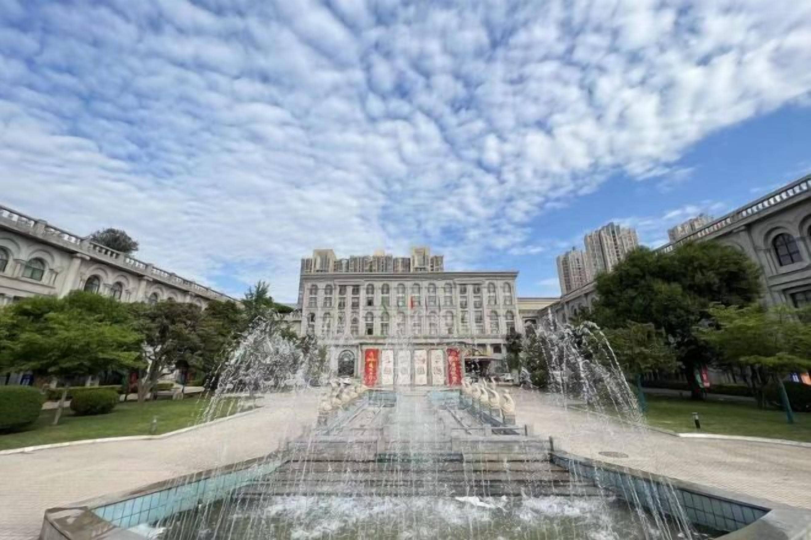 Un grande edificio con una fontana al centro, circondato da alberi e un cielo sereno.