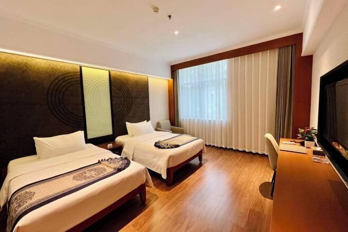 A hotel room with two beds, a desk, a television, and a large window with curtains.