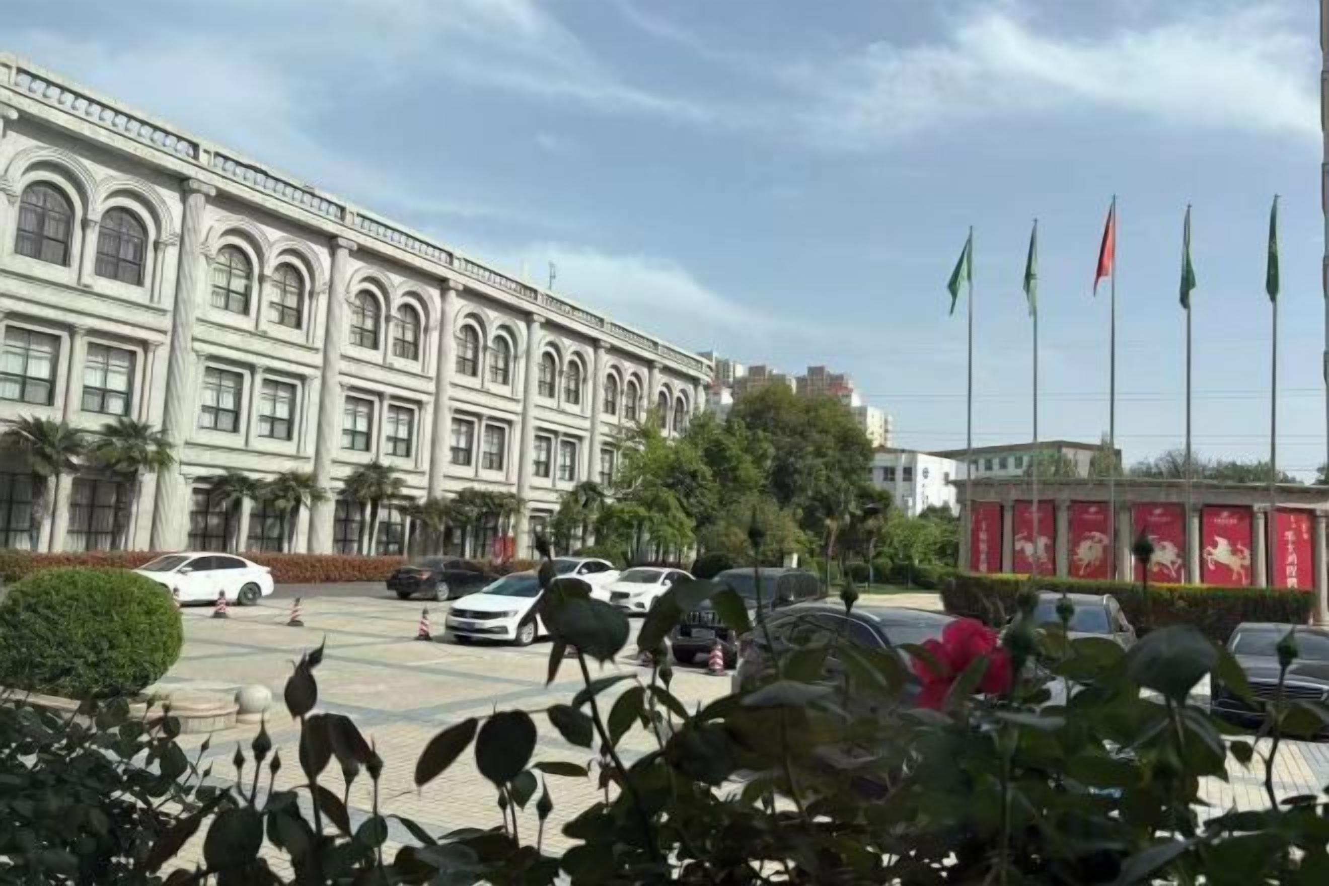 A building with a classical architectural style, a parking lot with several cars, and a red kiosk with flags.