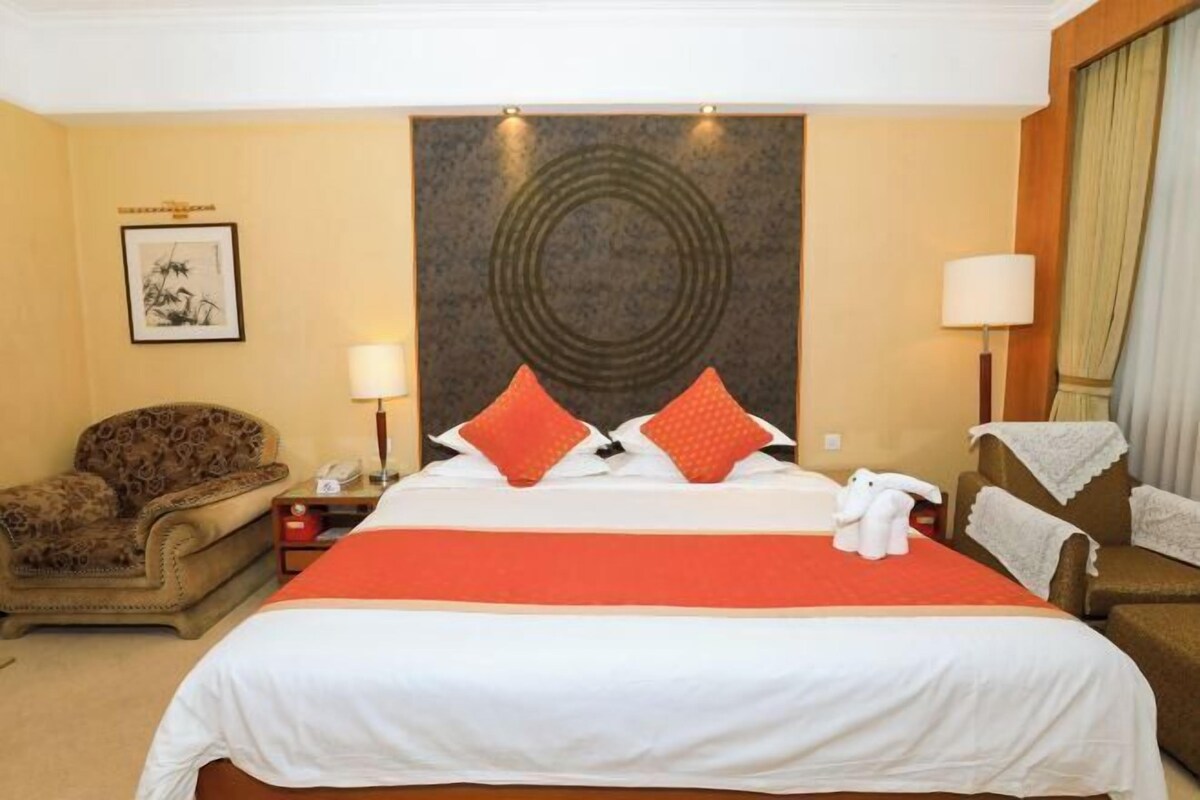 A hotel room with a large bed, two pillows, a nightstand, a lamp, a chair, and a wall art piece.