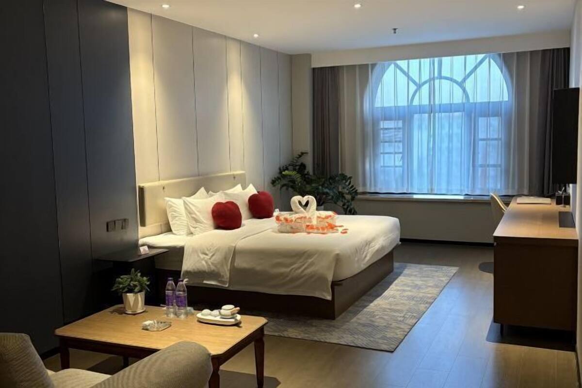 A modern hotel room with a large bed, a desk, a small table with a plant, and a large window with curtains.