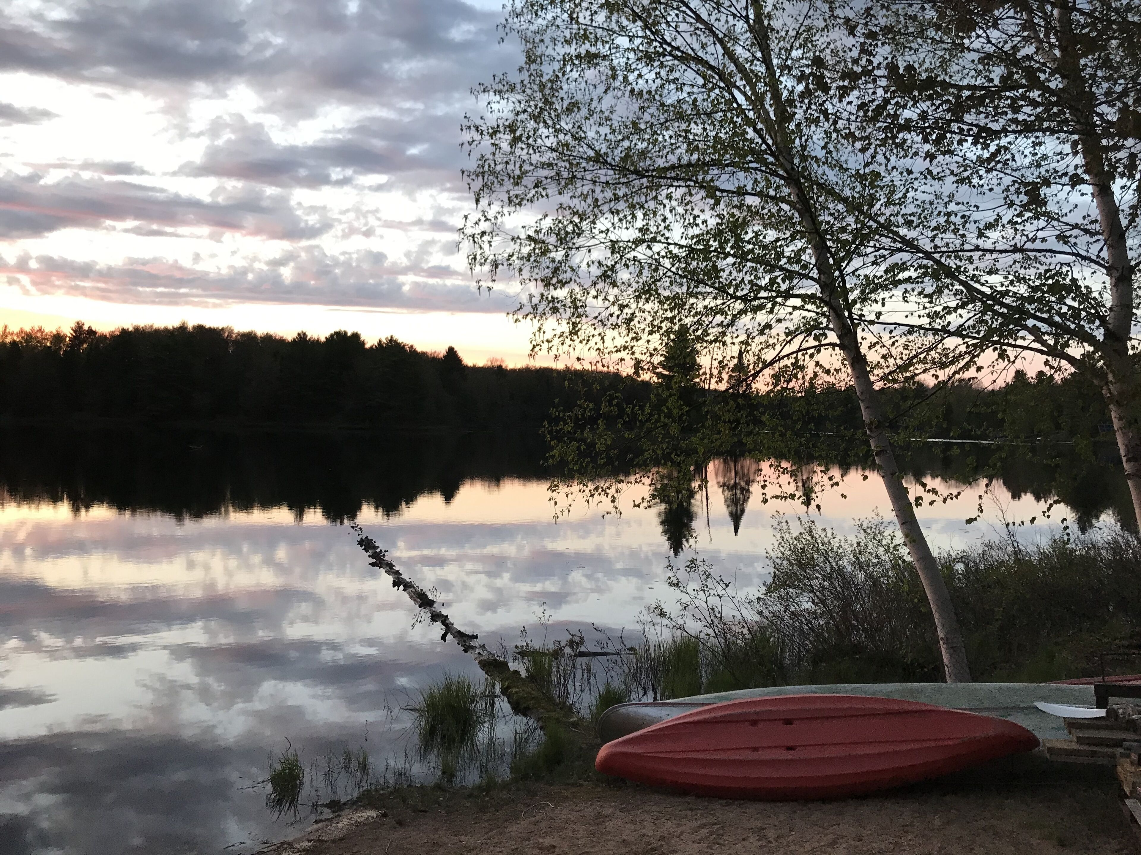 A calm lake at sunset with a red boat on the shore and trees reflecting in the water.