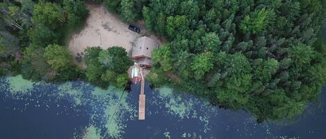 Vista aérea de una cabaña apartada rodeada de un bosque denso, conectada a un muelle que se extiende sobre un lago con vegetación flotante.