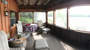 Una cabaña con un ventanal grande que da a un lago, una mesa con sillas y un pequeño rincón de estar con cojines.