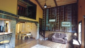 Una habitación amplia con techo de madera, pared de piedra, un sofá, una mesa de comedor y una escalera.