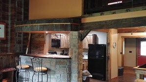 Una cocina rústica con una isla de piedra, taburetes de madera, un refrigerador negro y techo de madera.