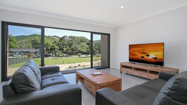 A modern living room with a large TV, leather sofas, a wooden coffee table, and a sliding glass door leading to a backyard. - BrightAway - Located in Bright, Victoria (BRIGHT)